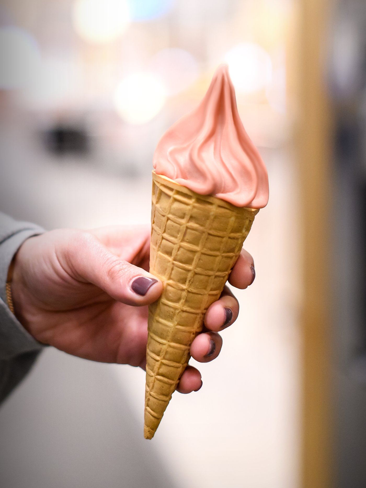 A hand of a person wearing a grey top and purple nail varnish holding a cone with vegan strawberry soft ice.