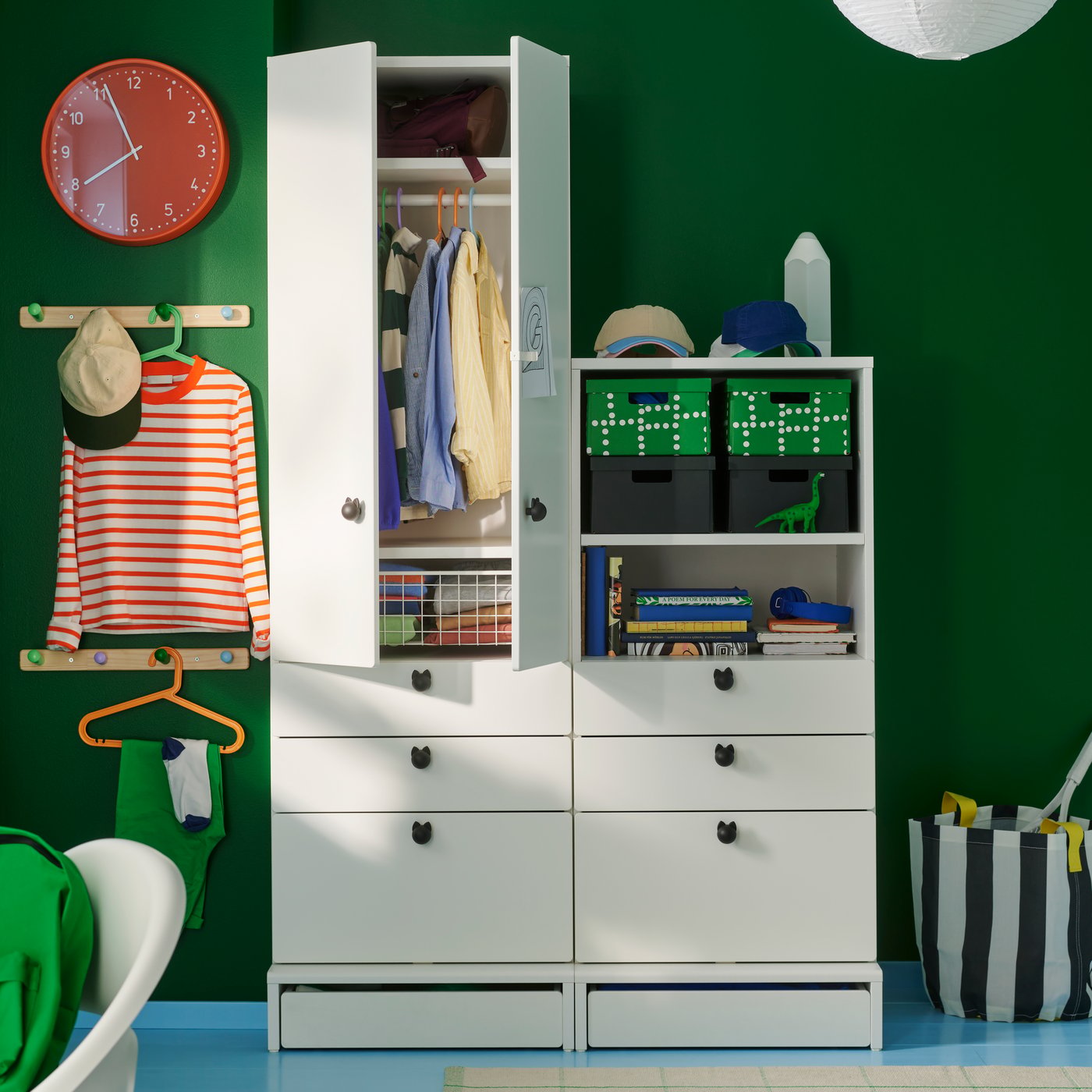 A BONDIS wall clock in red, mounted on a green wall beside a white wardrobe combination in a children’s bedroom with clothes.