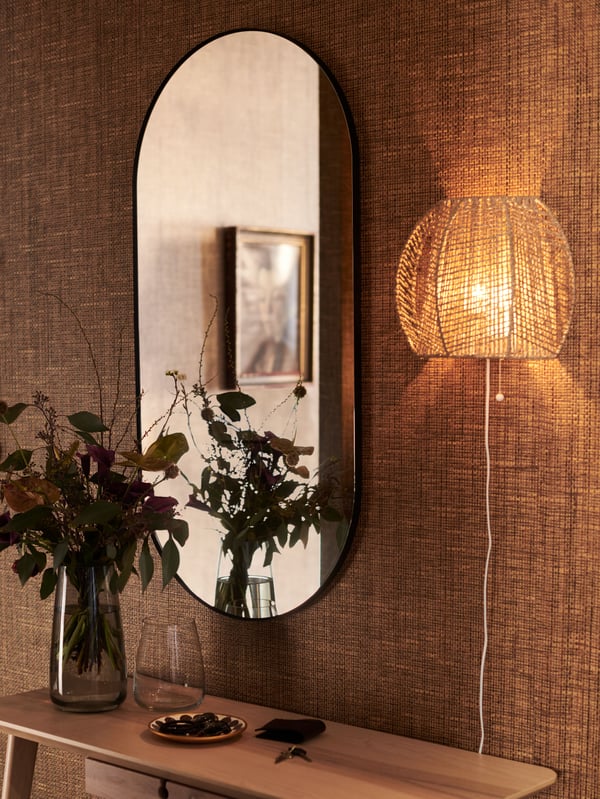 A MÅNALG wall lamp is mounted on a wall near a LINDBYN mirror and above a LISABO desk with flowers in a vase on top of it.