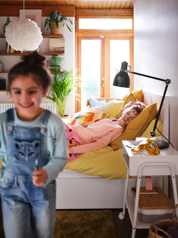 En lille piger løber grinende væk fra en seng, hvor der ligger en ung pige og sover.