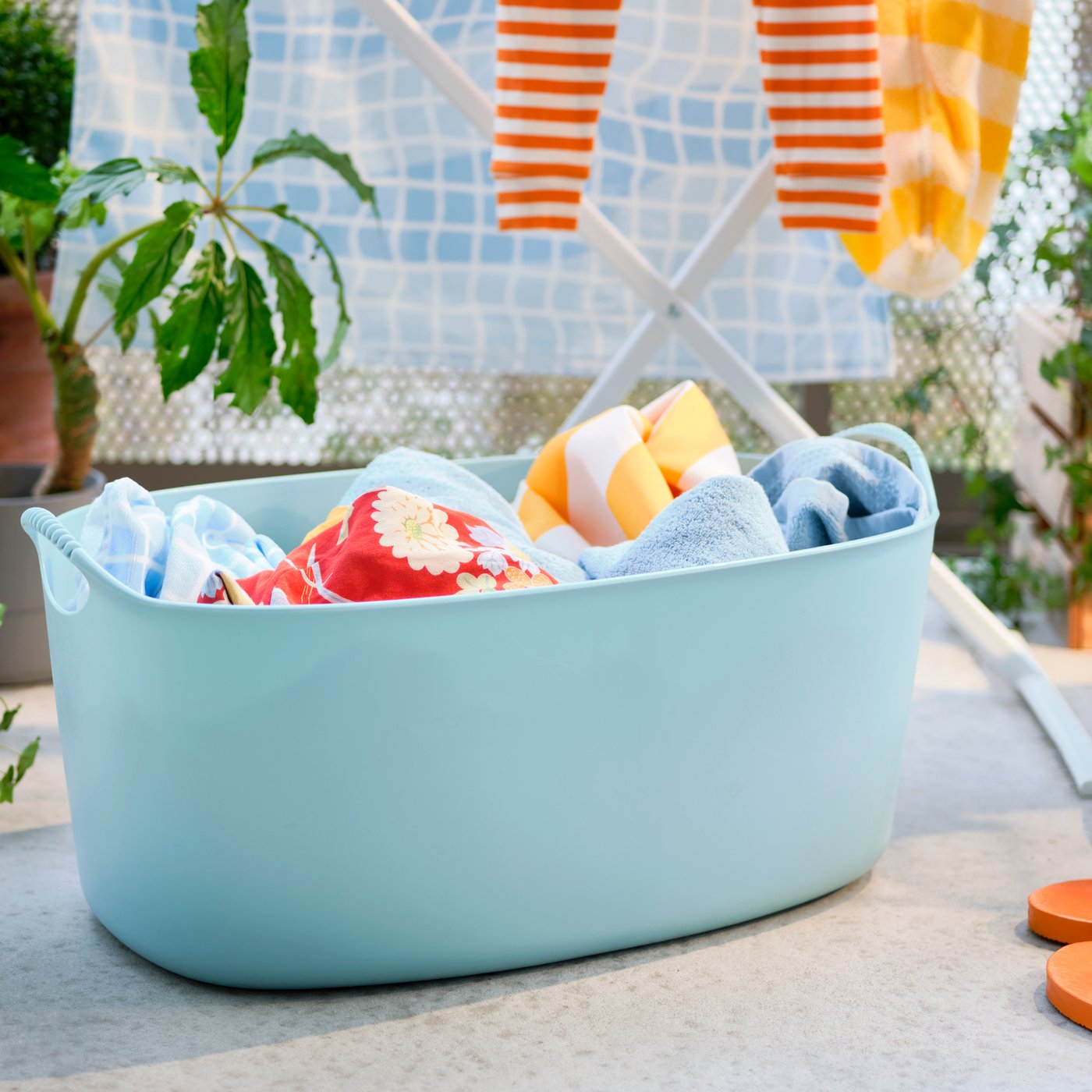 Su un balcone decorato con piante, portabiancheria flessibile TORKIS blu con biancheria, stendibiancheria e pouf SANDARED.