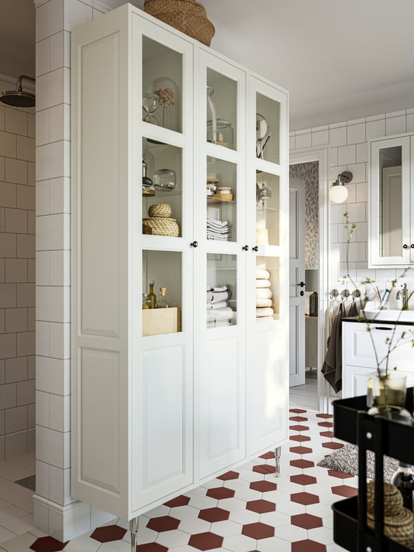 Three white TÄNNFORSEN high cabinets with doors, on two TÄNNFORSEN stainless steel legs, in a white tiled bathroom.