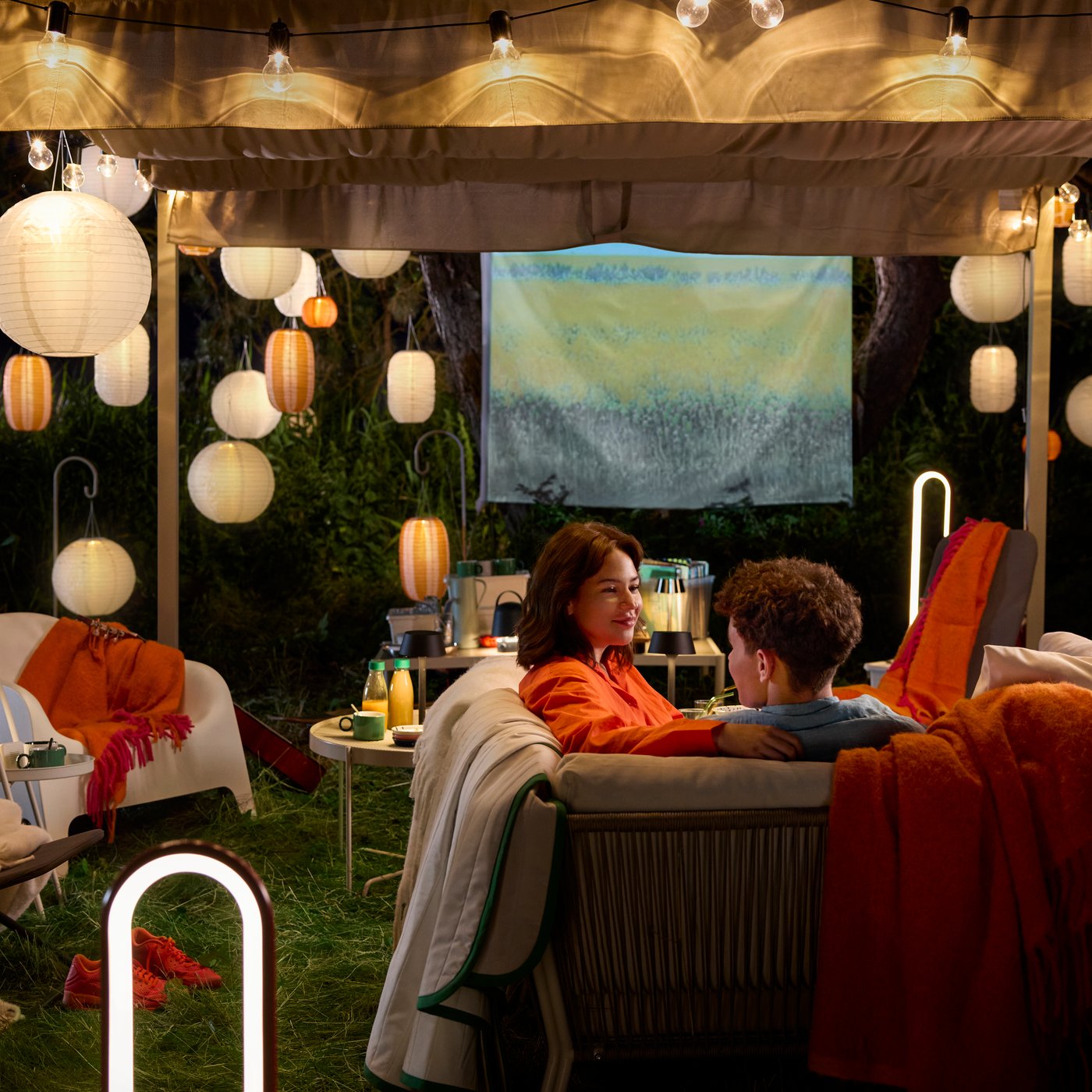 An outdoor seating area adorned with orange and white SOLVINDEN pendant lamps. Two young people in cosy blankets and cushions.
