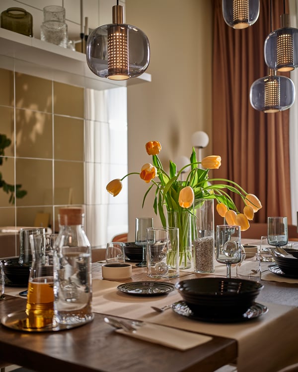A dining table is set with black bowls and plates, glassware and vase with tulips, with HÖGVIND pendant lamps hanging above.
