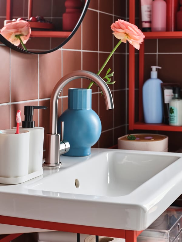 A white wash-basin with a soap dispenser, a toothbrush holder and a tray in white and a blue vase with a pink rose inside.