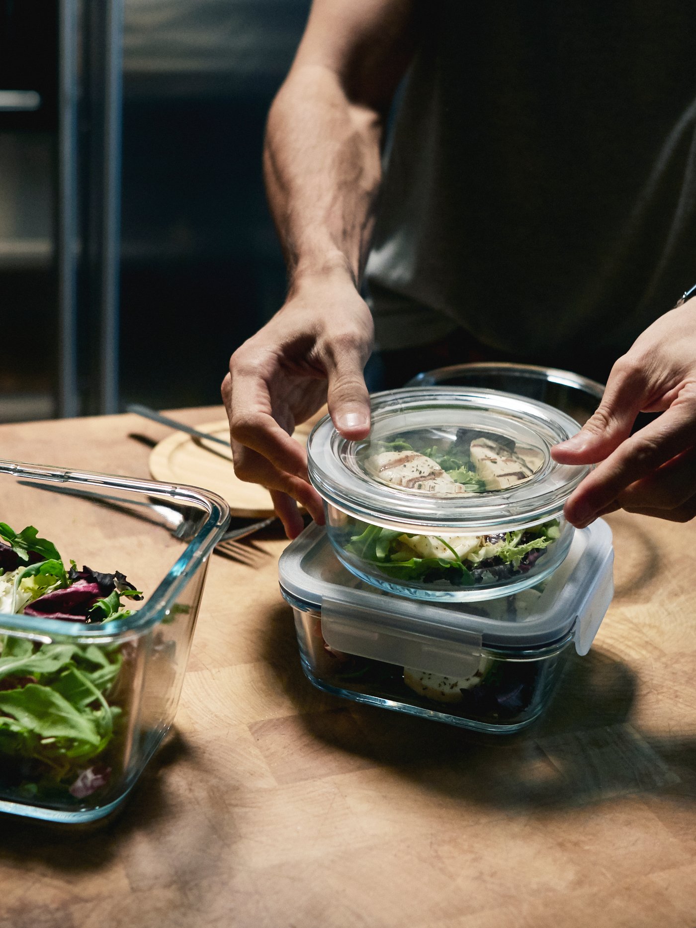A person in a kitchen putting the lid on a glass container of food. On the same benchtop are two other full food containers.