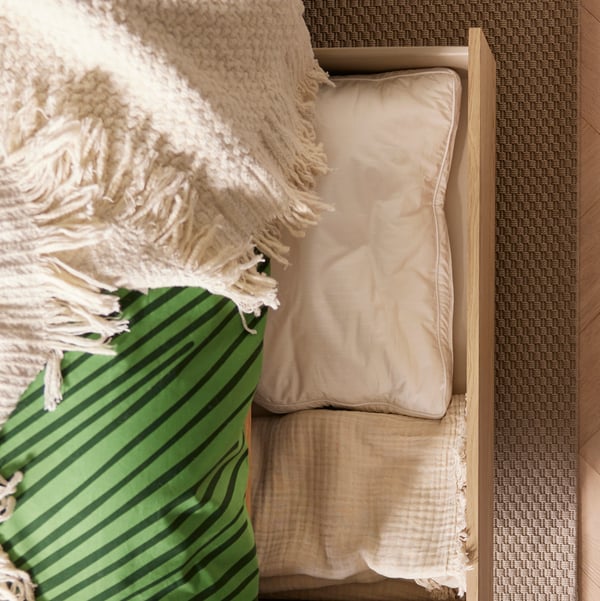 A LAPPTÅG throw lies on a bed. In one of the drawers of the bed, there is a cushion and a VALLKRASSING throw.