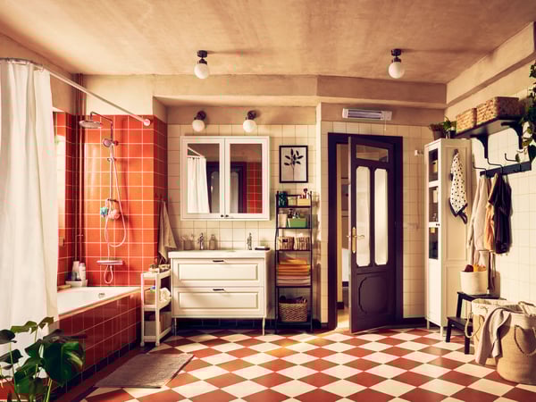 Salle de bain vintage avec des murs de couleur beige/terre cuite et un sol en damier dans les tons crème et terre cuite. Sur la gauche, une douche avec un rideau blanc et du carrelage rouge. Au centre, un meuble lavabo blanc avec miroir et appliques. À droite, une porte en bois partiellement ouverte donne sur une autre pièce. Une petite étagère contient des serviettes et des articles de toilette. En face de la porte, un portant avec des vêtements et des chapeaux. 