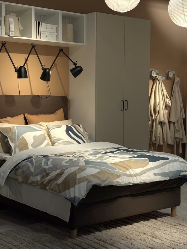 Modernt sovrum med bäddad HEMFJÄLLET säng i beige och grått, tre svarta vägglampor, hylla med böcker ovanför sängen, garderob och krokar med morgonrockar. Varm och inbjudande känsla.