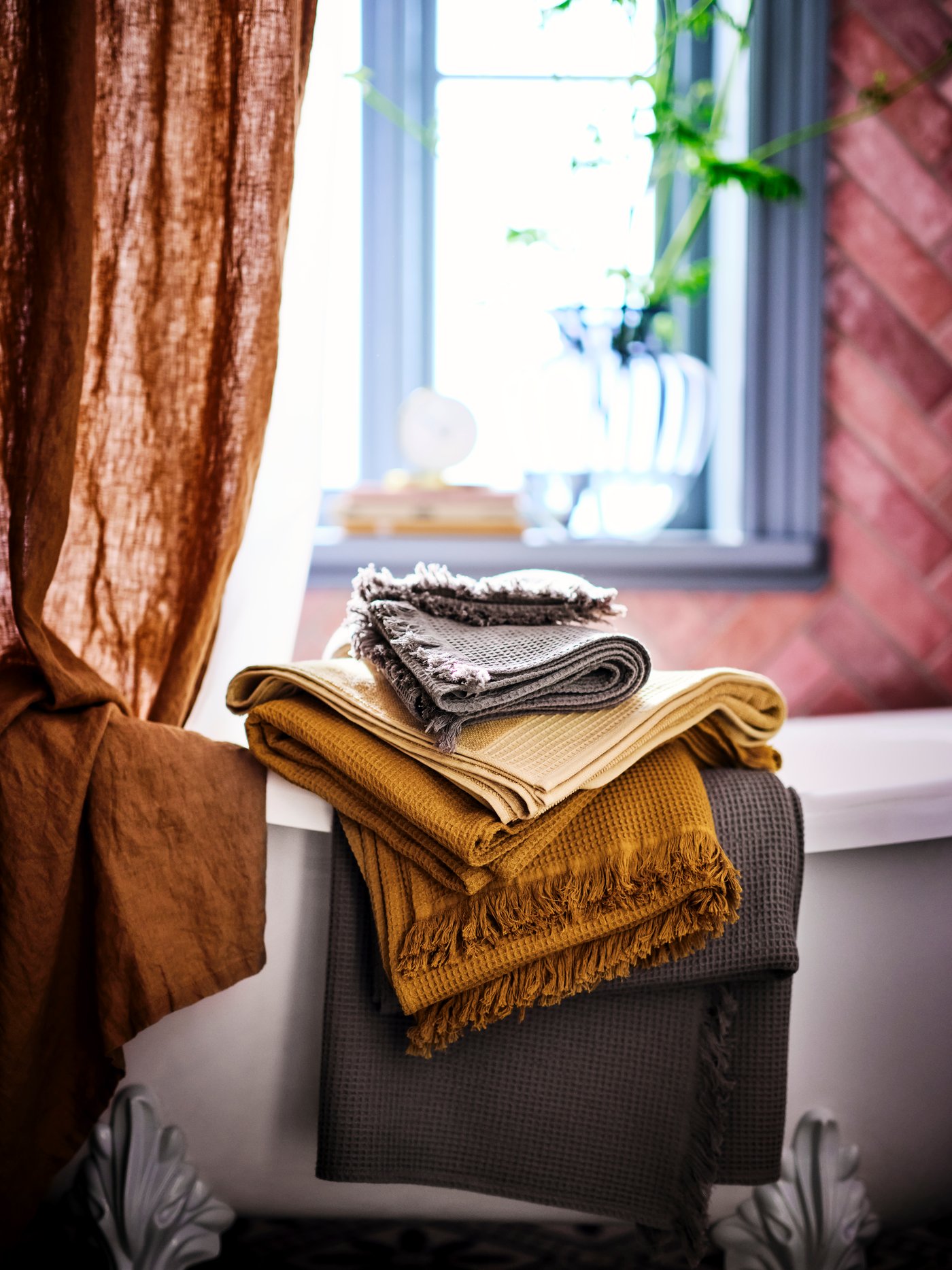 A light grey/brown VALLASÅN bath towel with others and a light yellow VINARN bath towel in a pile on the corner of a bathtub.