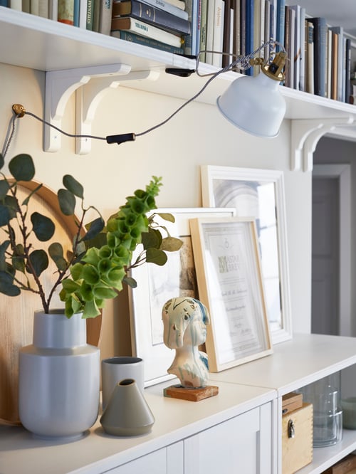 White BERGSHULT wall shelves mounted next to each other for a continuous bookshelf at the top of a wall above a cabinet.