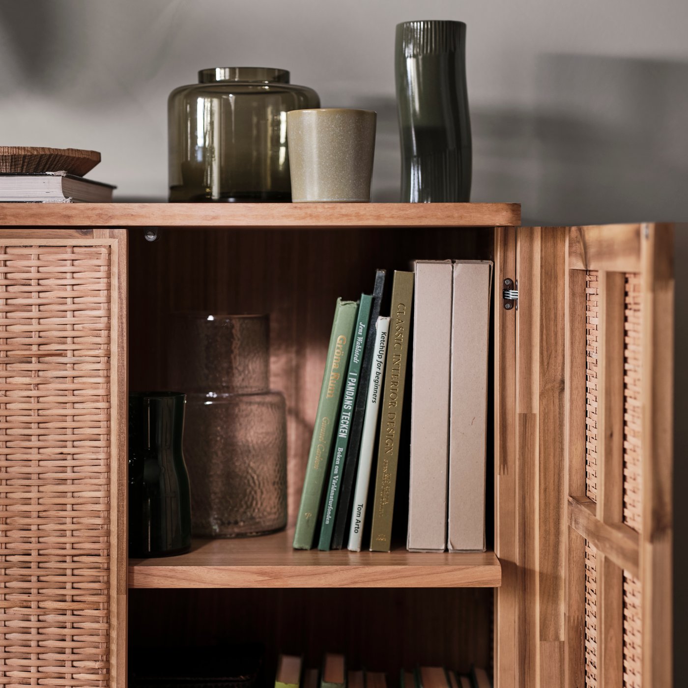 Träskåp med rottingdörrar, ena dörren öppen och visar hyllor med böcker och en KONSTFULL vas. Fler vaser står ovanpå skåpet.