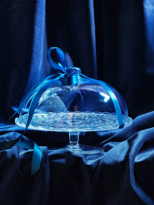 Close-up view of dining area with blue curtains, blue household textiles, glass serving stand with lid with blue ribbon.