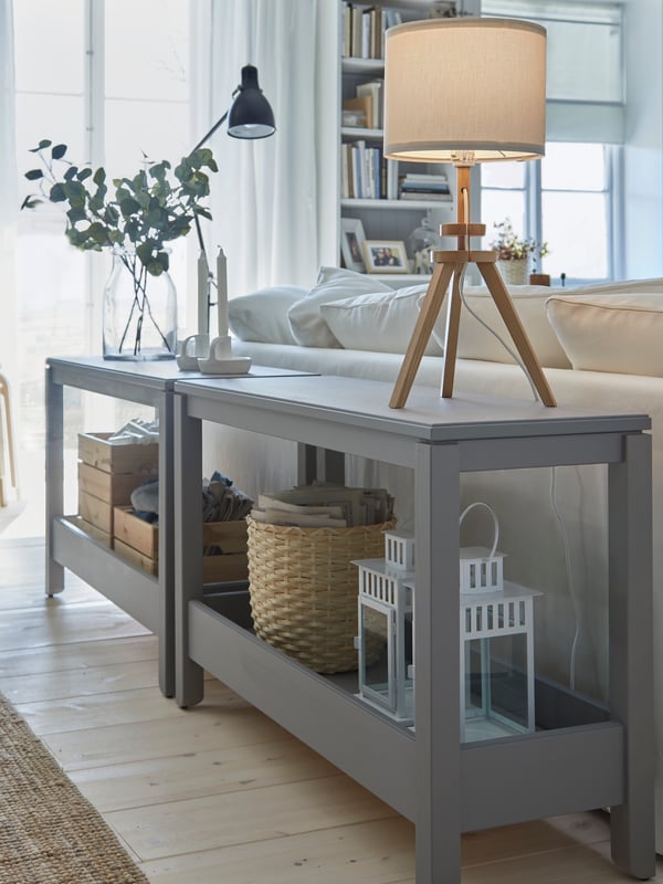 Two grey console tables are placed behind a sofa and serve as decorative room dividers. Two table lamps are placed on top.