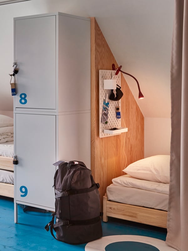 Glimpses of two hostel beds, separated by a wall and storage lockers, in the front there’s a black backpack.