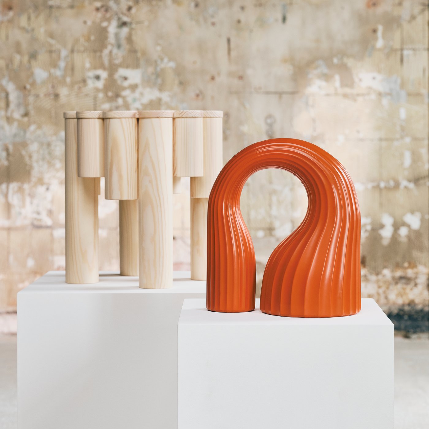 Close up of an orange sculpture and un-treated wooden stool placed on white pedestals in a rough gallery style studio.