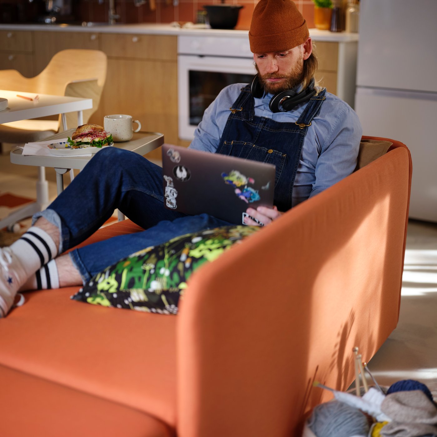 A person is sitting comfortably in a bright orange LINDÅKRA 2-seat sofa-bed with legs up and a laptop in their lap.