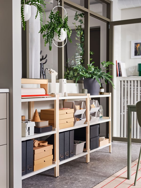 Three EKENABBEN open shelving units with some VATTENDRÅG boxes with lid and other things on them stand under some windows.