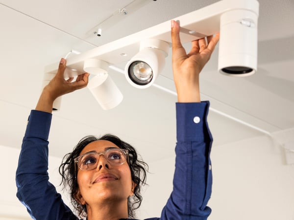 A woman in a blue shirt and glasses looks up while installing a white track lighting fixture on a white ceiling.