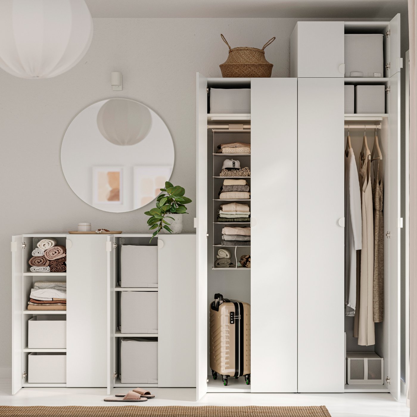 A minimalist white wardrobe and storage cabinet with open doors showing neatly organized shelves, hanging clothes, storage bins, folded towels, and a small suitcase, with a round mirror and plant on top.