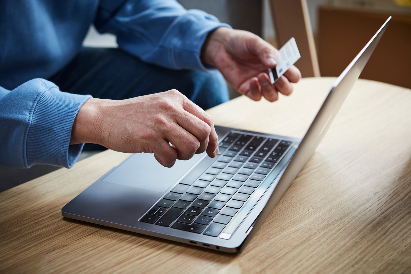 Personne portant un pull bleu utilise un ordinateur portable sur une table en bois et tient une carte de crédit dans une main, l’autre main étant posée sur le clavier.
