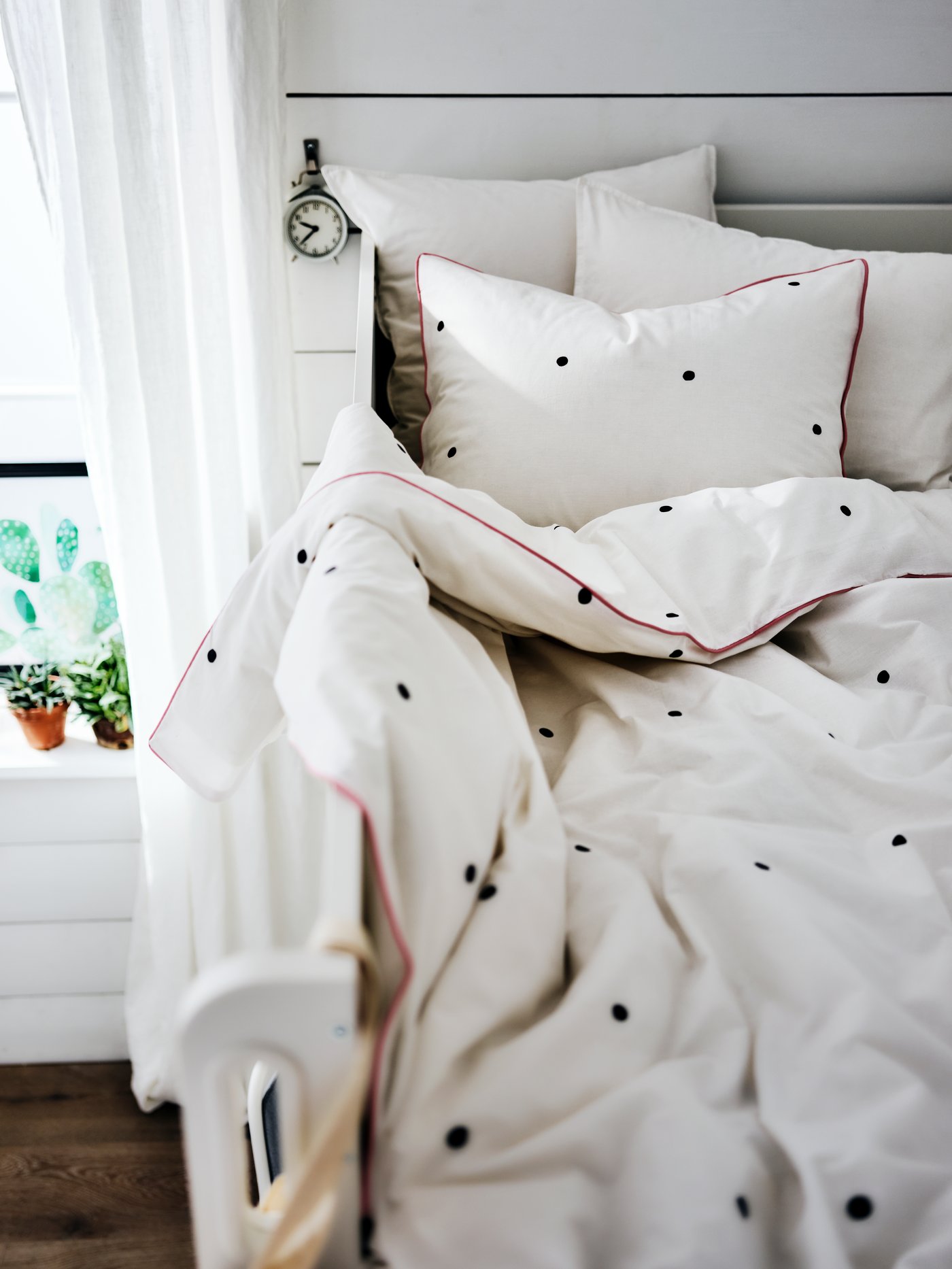 The top of a loft bed by a window with bedding that’s covered in white bed linen with a black spotted detail and a red trim.