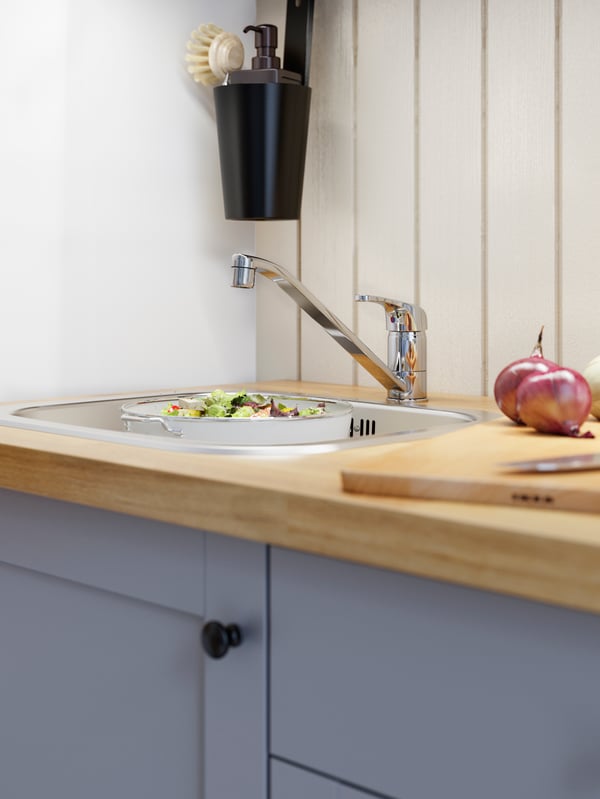 A wood worktop, grey kitchen fronts, an inset sink, a chrome-plated kitchen mixer tap and salad greens in a colander.