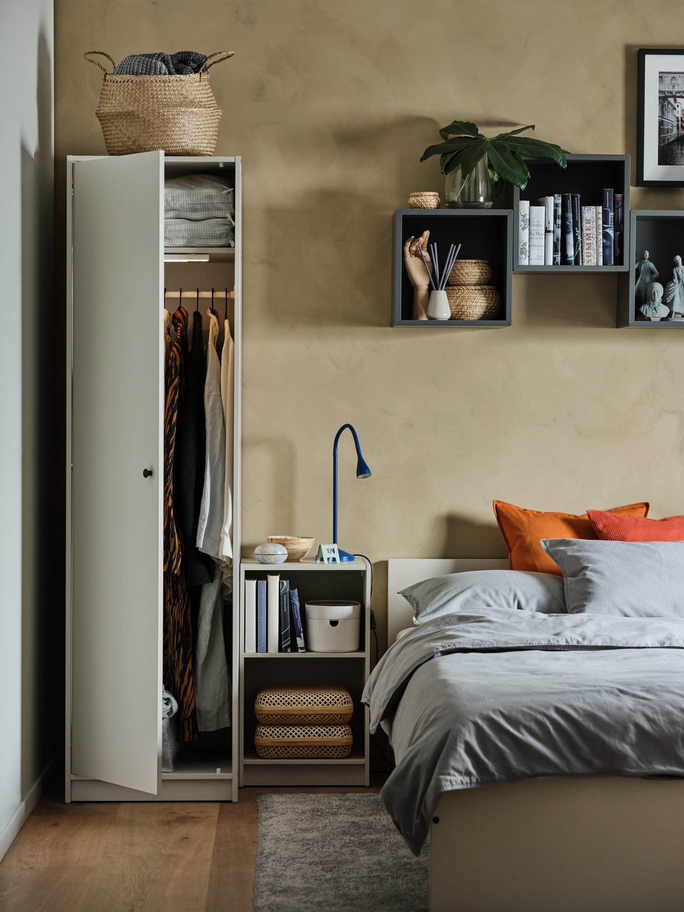 In a Bedroom there is a white cabinet with one door open on the left. Besides the cabinet there is a white Bed with grey bedding and over it there are pictures.