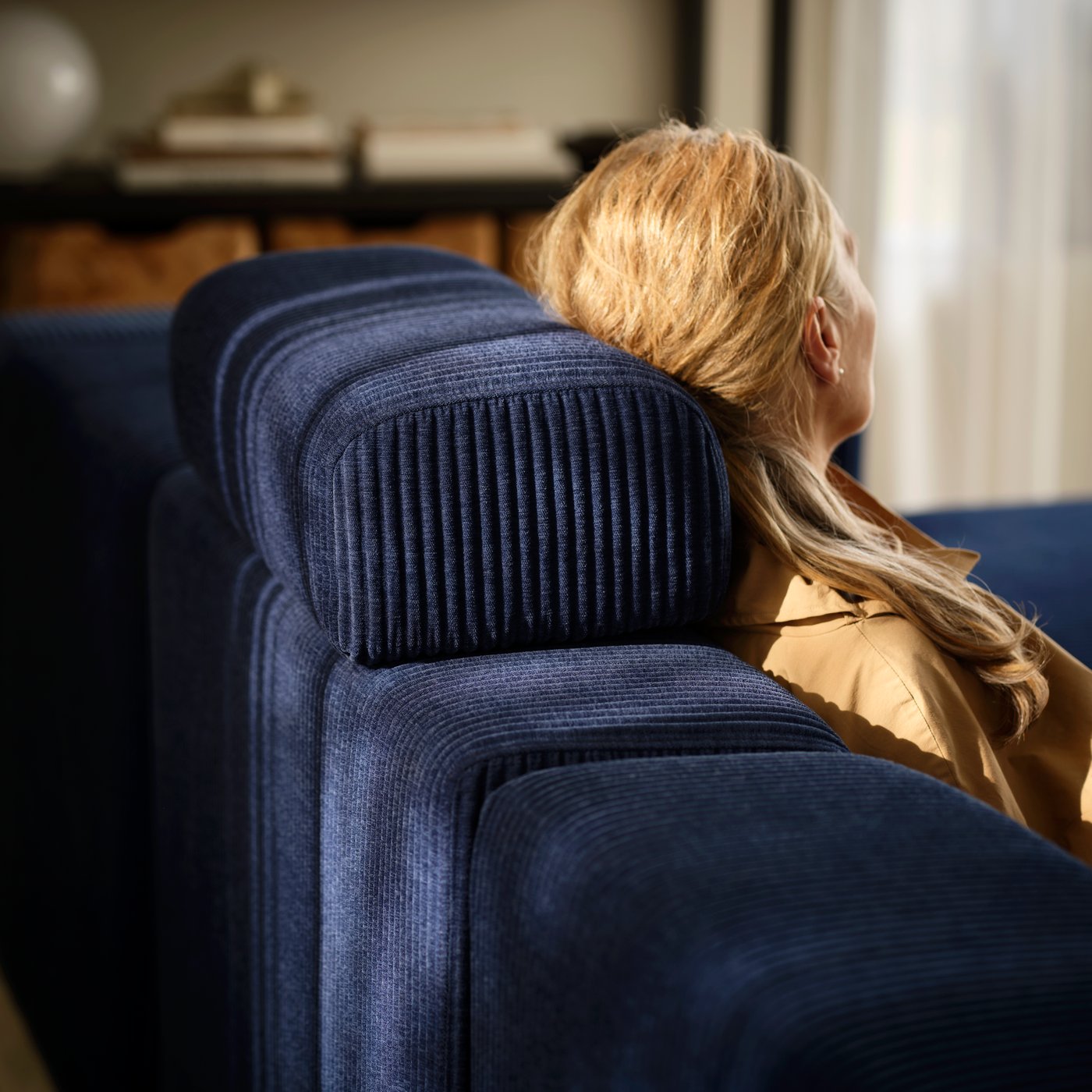 A woman leans back in a dark-blue JÄTTEBO four-seat sofa, her head against a loose headrest placed on top of the backrest.
