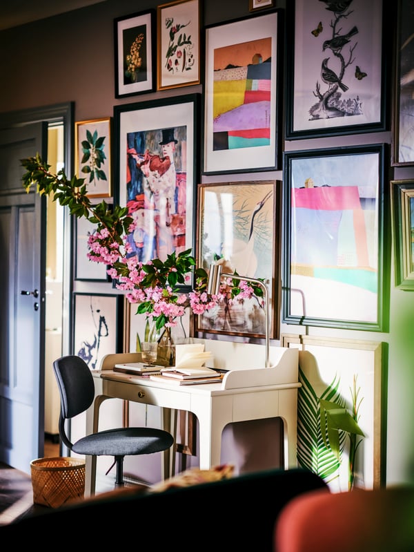 A BLECKBERGET swivel chair and a LOMMARP desk with a vase of flowers and notebooks on top against a wall of framed art.