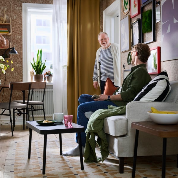 SONHULT nest of tables in black, placed on patterned rug in living room with sofa, wall art, curtains and two people.