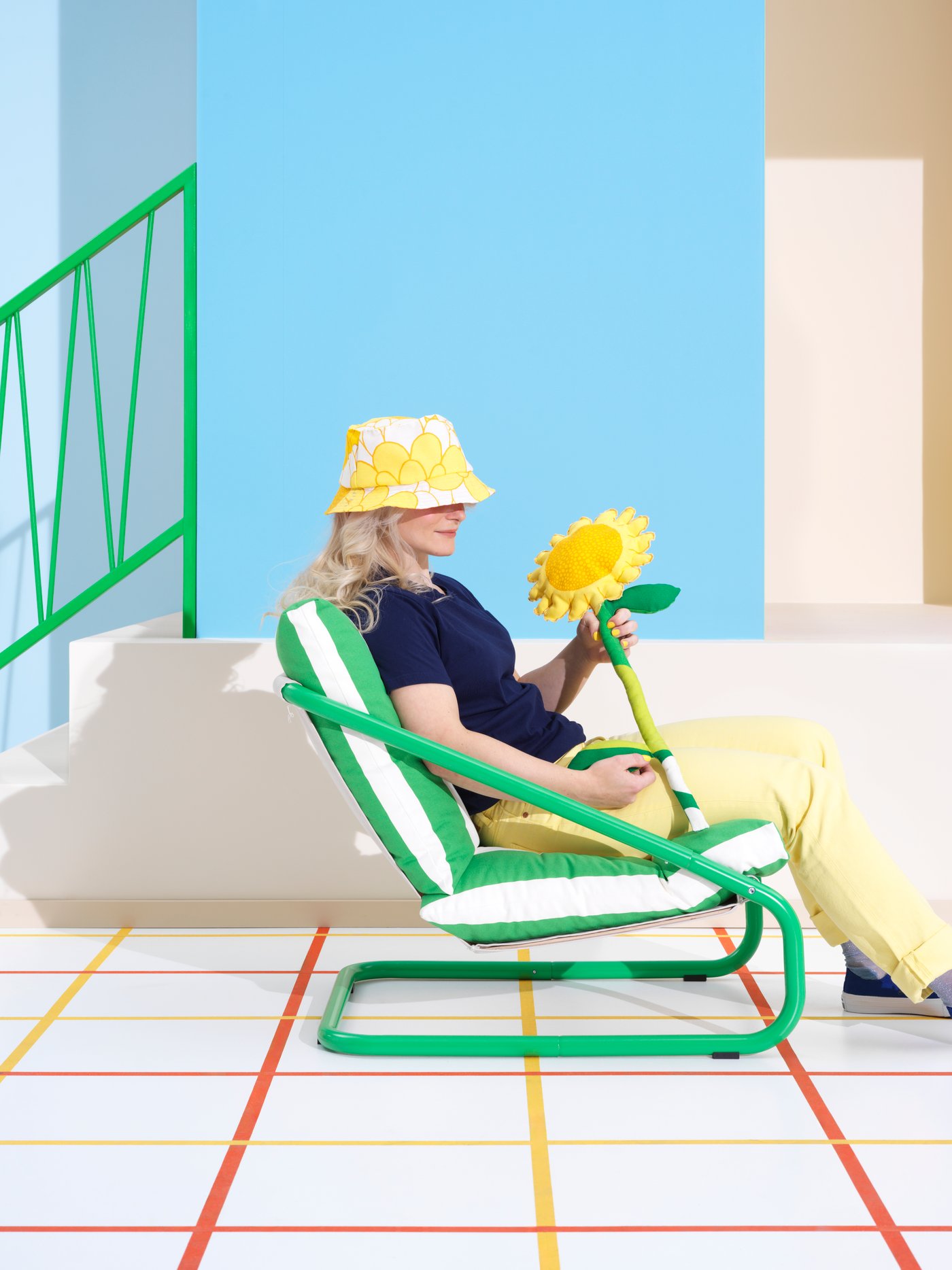 A girl in a yellow floral hat sits in a green striped ÖNNESTAD armchair in a bright living room, holding a fabric sunflower.