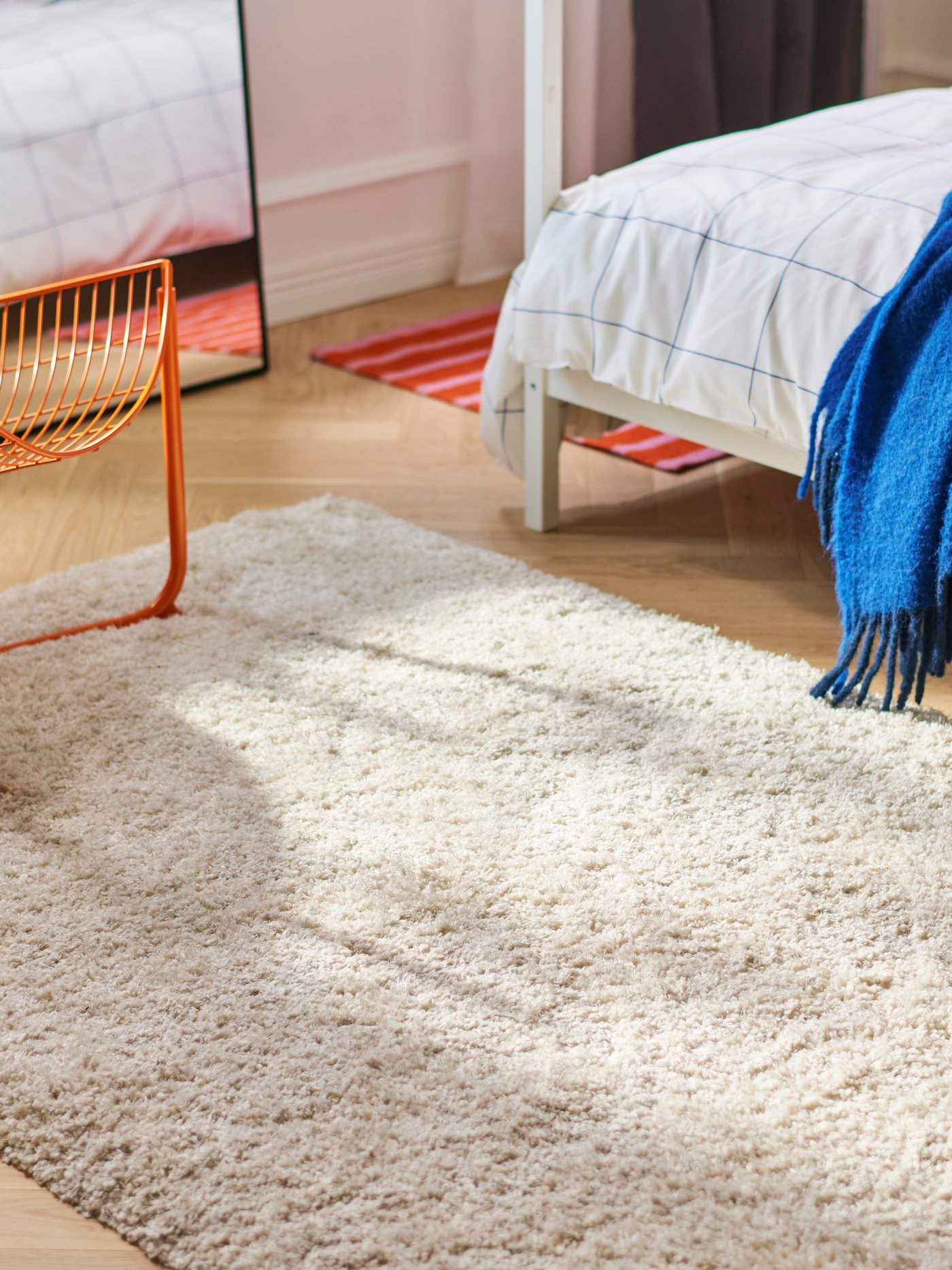 A cream-colored shaggy rug lies on the floor in a bright bedroom, beside an orange wire chair and a bed with a blue throw.