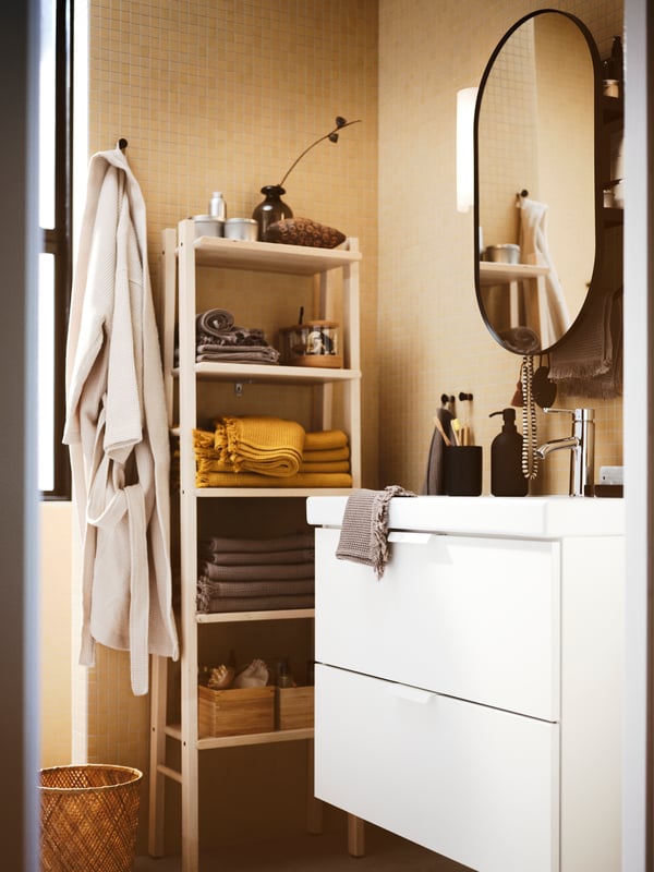 A bathroom with a mirror above a white GODMORGON wash-stand with two drawers, a wooden storage unit, towels and toiletries.