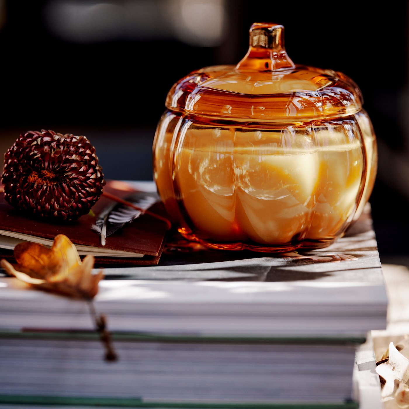 Una vela perfumada HÖSTAGILLE en un vaso con forma de calabaza con tapa, sobre un montón de libros y hojas secas.