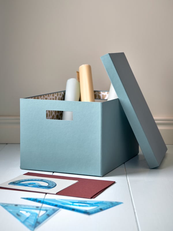 A paper storage box in blue holding rolls of paper, placed on a white floor and its lid leaning against the side of the box.