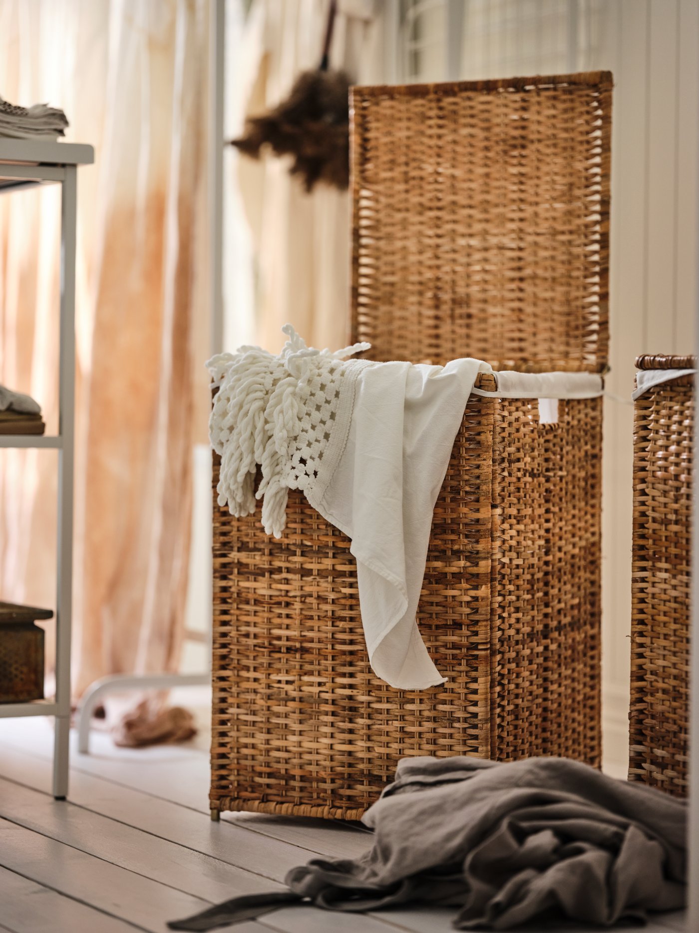 A room with white, wooden floor and walls. A BRANÄS rattan laundry basket, its lid upright, is overflowing with textiles.