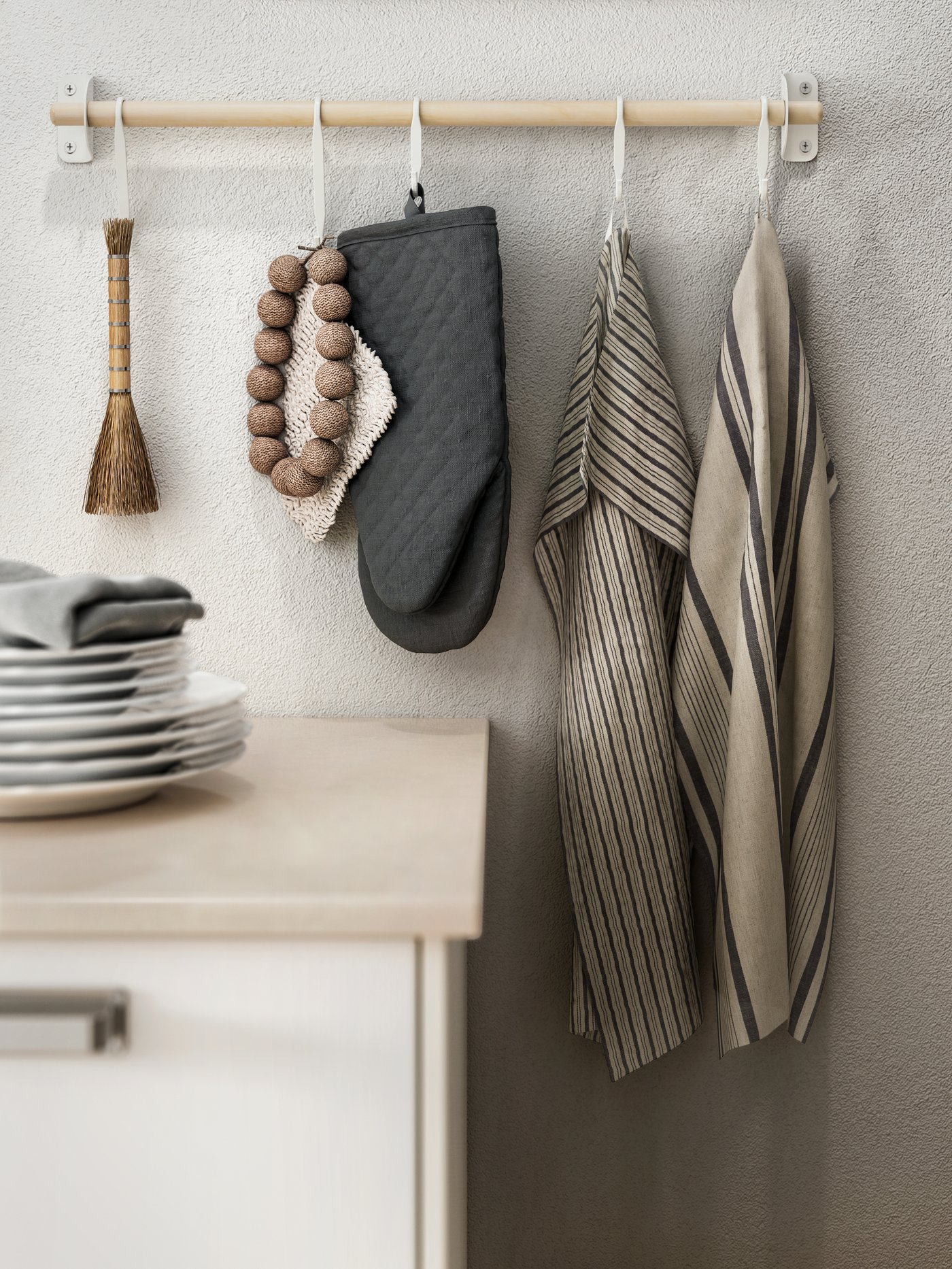 Tyč NEREBY na béžové stěně nad kuchyňskou linkou s talíři. Vedle šedé chňapky MARIATHERES visí dvě utěrky MARIATHERES.