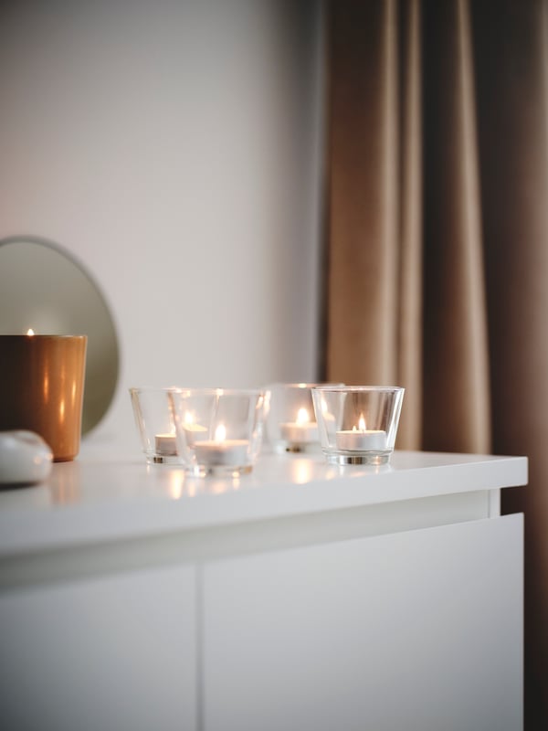 Four round GALEJ tealight holders in clear glass with burning candles, neatly grouped together on a white chest of drawers.