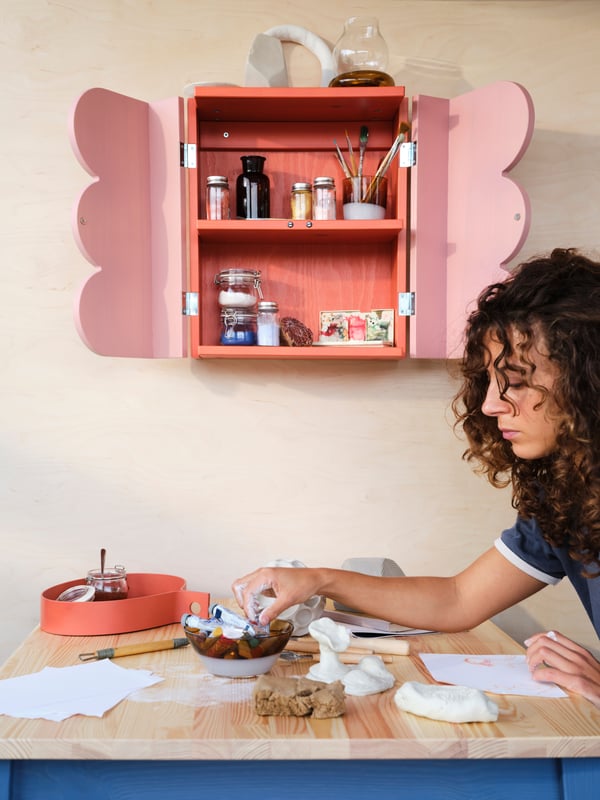 A person by a table picks up things from a bowl. An OMMJÄNGE cabinet with 2 doors and 1 shelf sits on the wall behind.