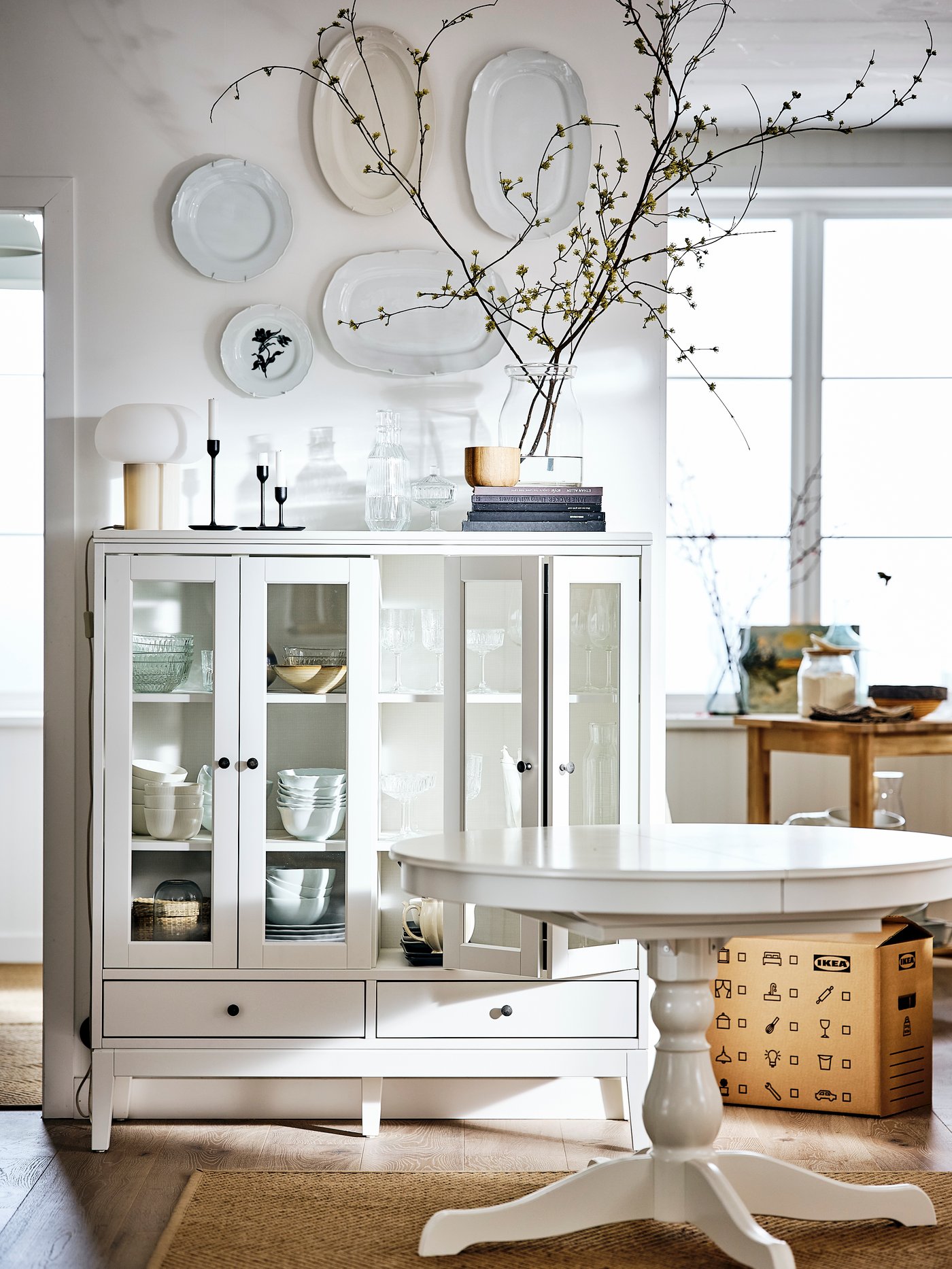 Salón con una mesa extensible blanca, una alfombra lisa, un armario con puertas de vidrio plegables, una lámpara de mesa y un jarrón.