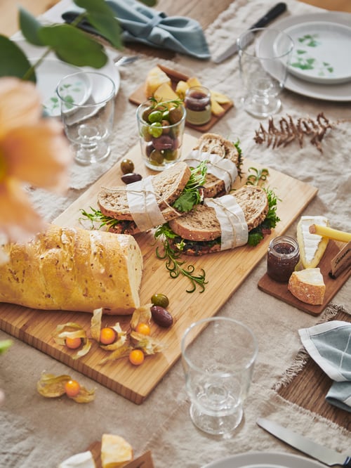 Dining table covered with a beige tablecloth, featuring several foods on a chopping board next to a goblet and a side plate.