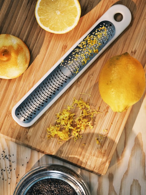 Overhead shot of an UPPFYLLD grater with lemon zest on its grates, a sliced lemon and a whole lemon on a chopping board.