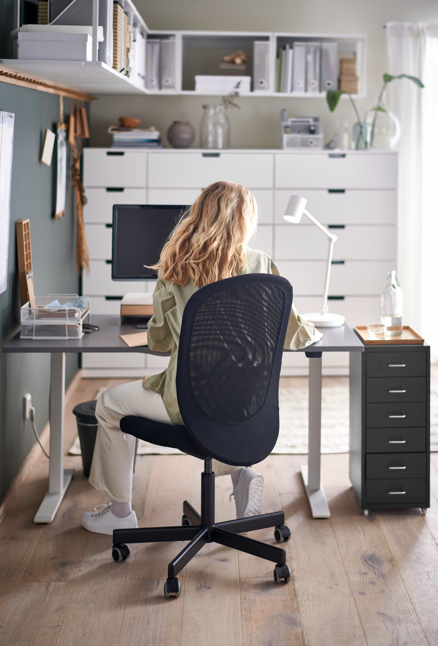 A person is sitting on a black FLINTAN office chair at her small home workspace. Next to the desk is a black HELMER drawer unit.