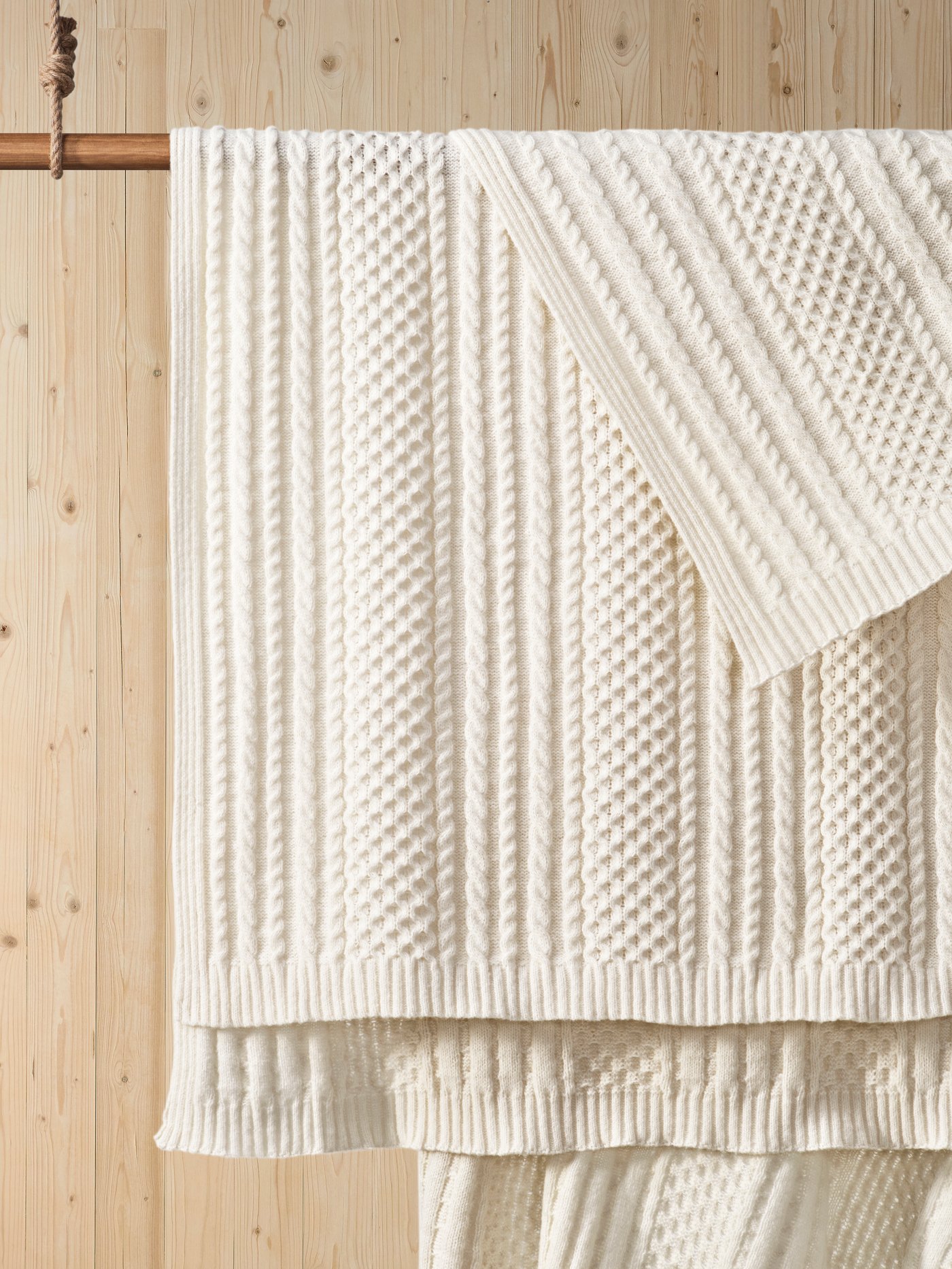 Cream-colored knitted throw blanket with a textured cable pattern draped over a wooden hanging rod.
