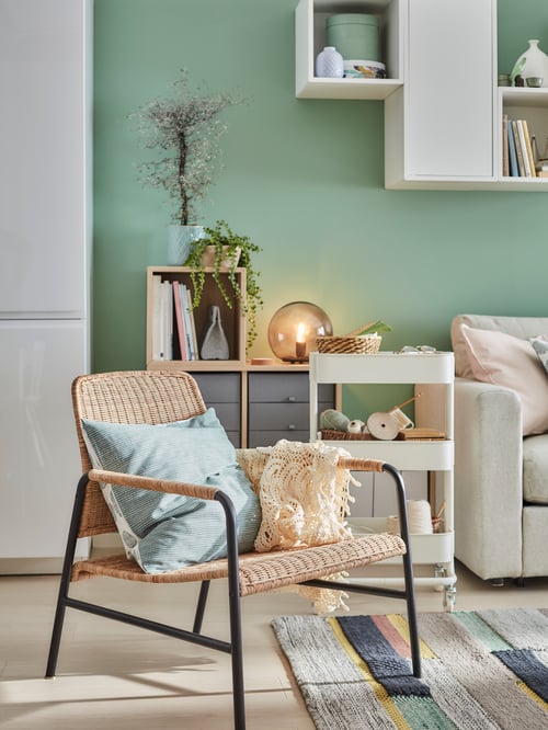 Living room with green wall, armchair, sofa-bed, trolley, table lamp, low pile rug, and wall-mounted cabinet.