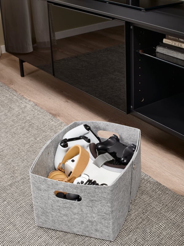 Une boîte de rangement en feutre IKEA JÄGMÄSTARE grise remplie de plusieurs manettes de jeu, posée sur un tapis gris devant un meuble TV noir.