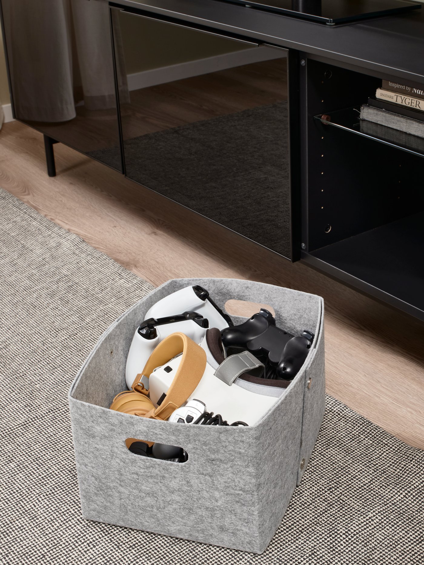 Een grijze IKEA JÄGMÄSTARE opbergdoos van vilt gevuld met verschillende gamecontrollers, geplaatst op een grijs vloerkleed voor een zwart tv-meubel.