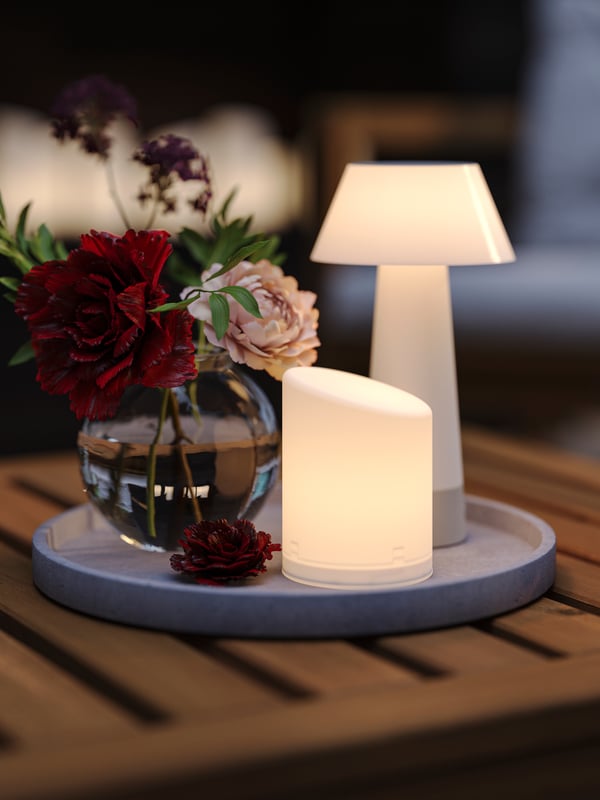A lit in/outdoor ÄDELLÖVSKOG LED block candle and a battery-operated table lamp on a light grey saucer on a wooden table.