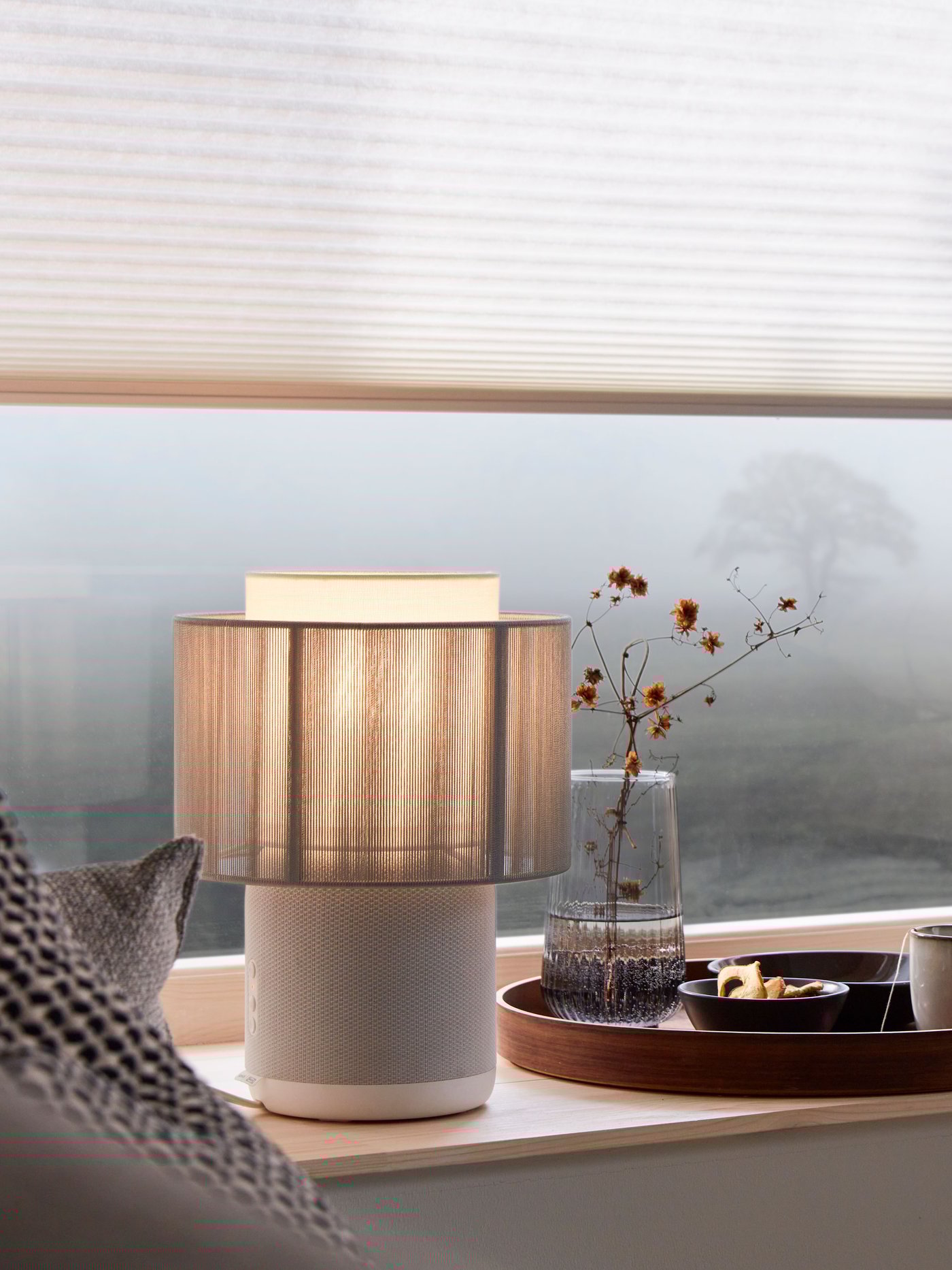 Eine SYMFONISK Tischleuchte WiFi-Speaker beleuchtet einen Fensterbereich. Vor dem Fenster ist eine nebelverhangene Landschaft zu sehen. Am Fenster hängt ausserdem eine PRAKTLYSING Wabenjalouise.